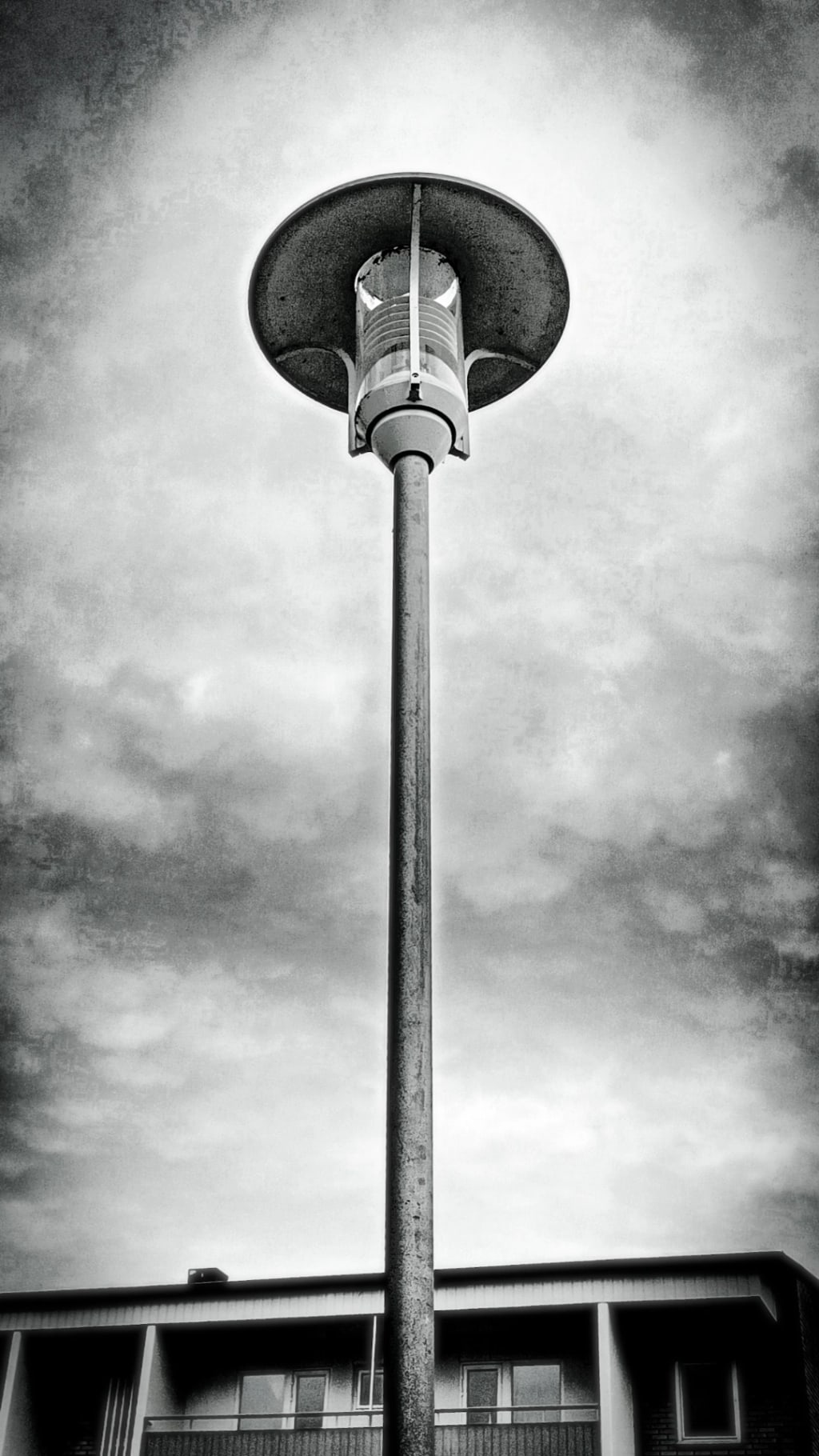A stark black-and-white view of a tall light pole against cloudy sky.